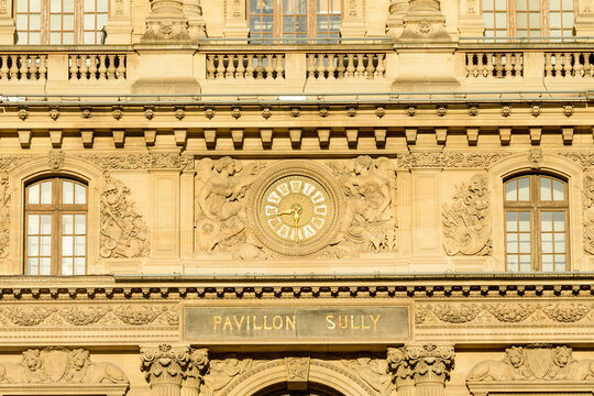Detailed frontal view of the Pavillon Sully facade at the Louvre in Paris, France, featuring a central clock surrounded by intricate classical stone bas-reliefs and ornate architectural details