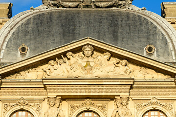 Obraz premium Close view of the ornate pediment on the Louvre palace in Paris, France, featuring detailed classical stone sculptures illuminated by warm sunlight. The historic reliefs and slate dome create a