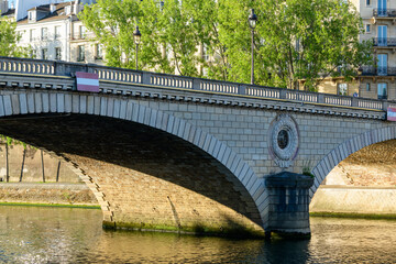 Fototapeta premium Sunlit view of Le Pont Louis-Philippe spanning the Seine River, with detailed masonry, ornate medallion, and leafy trees lining historic Parisian buildings in the background.