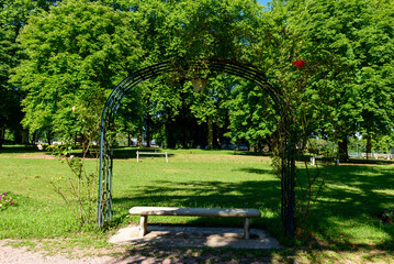 Obraz premium A rustic stone bench sits beneath a wrought iron arch entwined with climbing roses, surrounded by lush green grass and tall leafy trees in the sunlight at Saint-Honore-les-Bains.