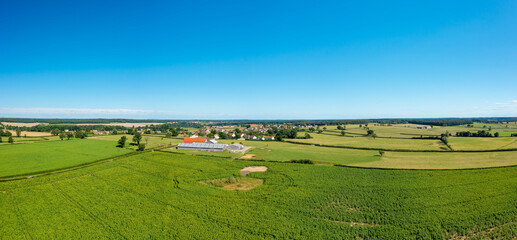 Wide panoramic aerial view of open green fields and agricultural buildings near Saint-Germain-Chassenay, under a vivid blue summer sky. The landscape features layered textures, rolling meadows, and a