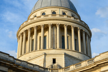 Low angle view of the Pantheon's neoclassical dome and colonnade in Paris, bathed in warm golden sunlight with clear architectural lines and soft blue sky.