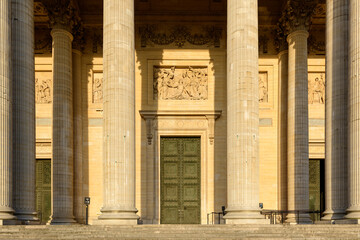 Obraz premium Close-up of the Pantheon's grand entrance in Paris, featuring tall neoclassical columns, ornate green doors, and sculpted stone reliefs illuminated by warm afternoon sunlight.
