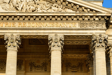 Fototapeta premium Close-up of ornate Corinthian columns and detailed stone pediment on the Pantheon in Paris, illuminated by golden sunlight with crisp shadows highlighting intricate carvings.
