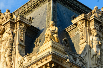 Fototapeta premium Golden evening light highlights a group of detailed stone sculptures and ornate reliefs at the corner rooftop of the Louvre palace in Paris, France. The renaissance architecture features intricate
