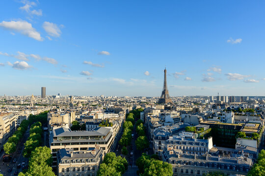 Expansive panoramic view over Paris featuring the Eiffel Tower rising above classic city rooftops and tree-lined avenues under a bright blue sky with scattered clouds.