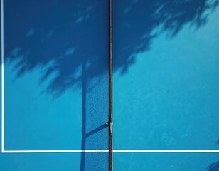 Blue paddle tennis court from above. Net divides the game surface. Tree shadows cast patterns on synthetic turf. Outdoor sport background for leisure activities.