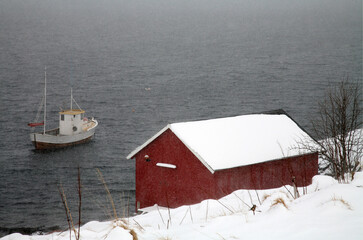 Fishing boat in winter during snowfall in front of a cabin on the fjord in Lofoten, Norway
