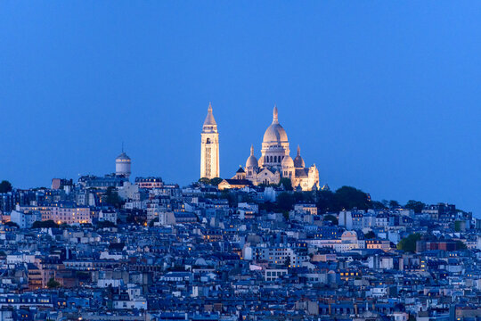 The illuminated domes and bell tower of Sacre Coeur Basilica rise above the densely packed rooftops of Montmartre at dusk, set against a vibrant blue evening sky.