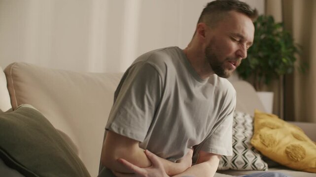 Man sitting on sofa at home, holding his abdomen and bending forward, suffering from acute stomach pain and digestive problem