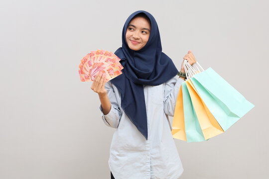 Portrait of a young Asian woman in hijab, happily holding Indonesian rupiah banknotes and shopping bags, standing isolated on a gray background.