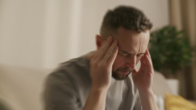 Distorted shot of man sitting on sofa at home, holding his temples with both hands and closing eyes due to intense headache