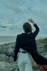 Fototapeta premium Coastline scene with figure in dark coat walking away on a rocky shore, wind blowing, white trousers, ocean backdrop and moody sky, suggesting adventure and contemplation.