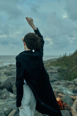 Obraz premium Woman stands on a rocky coast, wind in a dark jacket, raising a hand toward the sky as sea and clouds meet the horizon in a dramatic outdoor scene.