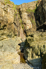 Steep stone cliffs at Cap de Carteret display layered textures and subtle green moss, illuminated by warm sunlight under a vivid blue sky. The rugged surface and vertical composition evoke a sense of