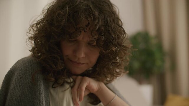 Curly-haired woman wrapped in blanket coughing into her fist and touching her forehead in pain while holding digital thermometer in hand during illness at home
