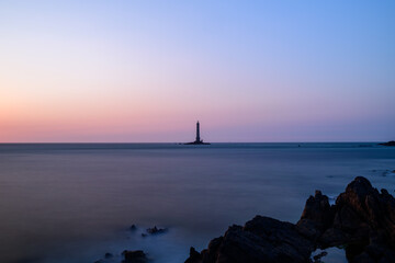 Long exposure at dusk reveals a tranquil sea with smooth water and pastel skies, featuring the Cap de la Hague lighthouse on a distant islet and rugged rocks in the foreground.
