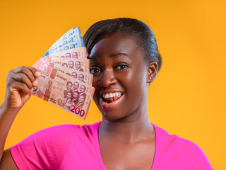 A woman with a joyful expression holds a fan of Ghanaian Cedi banknotes against a bright yellow background. She wears a pink shirt, showcasing the 200 and 100 Cedi notes with a celebratory smile.