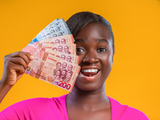 A joyful woman in pink holds a fan of Ghanaian Cedis against a bright yellow background. Her expression is one of pure excitement and financial triumph, celebrating wealth and success in Ghana.