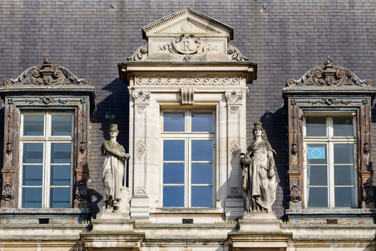 Two classical stone statues stand beside tall windows with intricate decorative frames on the historic slate facade of Hotel de Ville in Paris, illuminated by bright daylight.