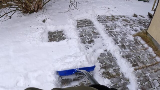 POV pushing a blue snow pusher along a cobblestone driveway edge beside a beige wall, scraping slush and powder into a berm to prevent refreeze and improve traction.