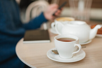 Fototapeta premium Aesthetic cafe moment: close-up of a white tea cup in focus with a person using a smartphone in the blurred background.