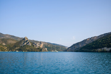 Fototapeta premium Bright turquoise waters of Lac de Castillon framed by rocky, forested hills and a distant sailboat beneath a clear blue sky.