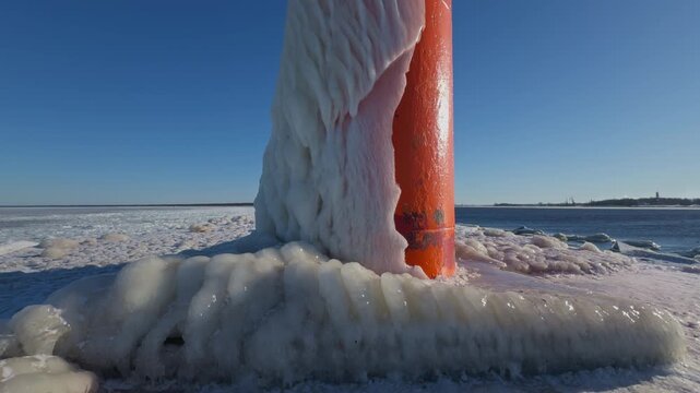 Ice surrounds orange tower base. Coastal horizon near frozen metallic structure with textured surface