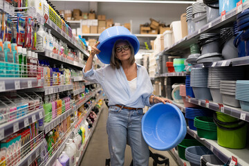 Woman playfully holds blue buckets on her head while shopping in a store aisle filled with cleaning supplies