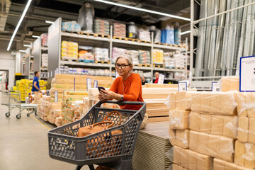 Woman using smartphone while resting in a hardware store aisle filled with materials and supplies