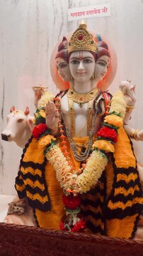 God Dattatreya depicted with three serene faces symbolizing Brahma, Vishnu, and Shiva, adorned with floral garlands and traditional attire, accompanied by a sacred cow inside a temple sanctum.