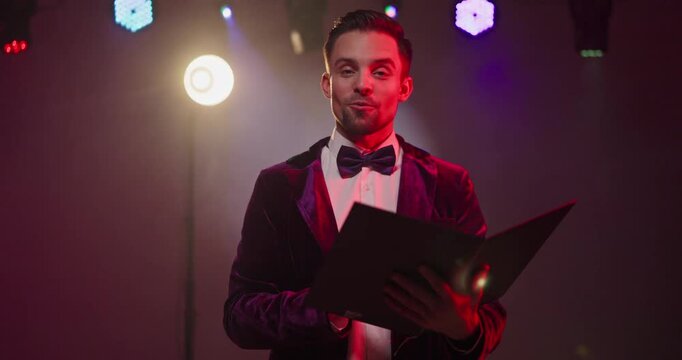 Presenter showman host on stage under spotlight. Smiling announcer and emcee with clipboard in bowtie jacket engages the audience amid lighting. Concept shows professional event hosting.