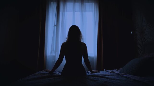 black silhouette upset unhappy woman sitting on bed dark room, mood depression sadness. Sad lonely Girl feels bad after party migraine suffers from headache. window curtains white fabric fly wind blow