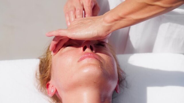 Therapist presses and glides on forehead and nose, oiled skin shines, stud earring and white linen show, veined forearms visible, daytime coastal spa Montenegro