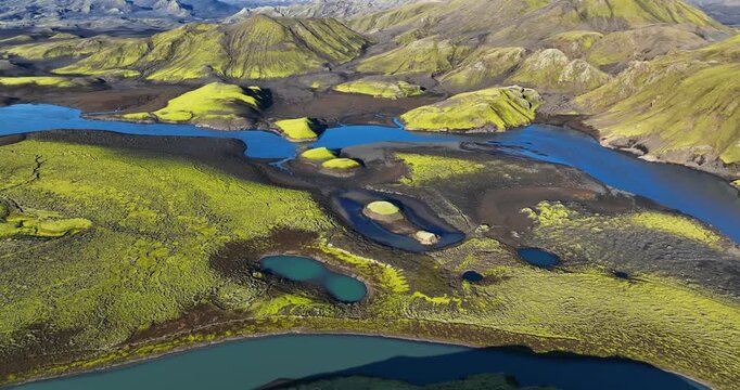Aerial view of Iceland highlands river wetlands with turquoise lagoons, mossy lava plains and black sand channels beneath rugged mountains in bright summer light.