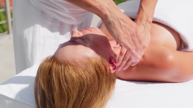 Daytime close up shows veined hands massaging neck, jawline, and trapezius on a white towel in Montenegro. Warm light and shallow depth of field suggest a spa.