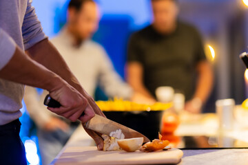 Obraz premium Close-up of a person slicing an onion on a cutting board with friends enjoying a gathering in the background