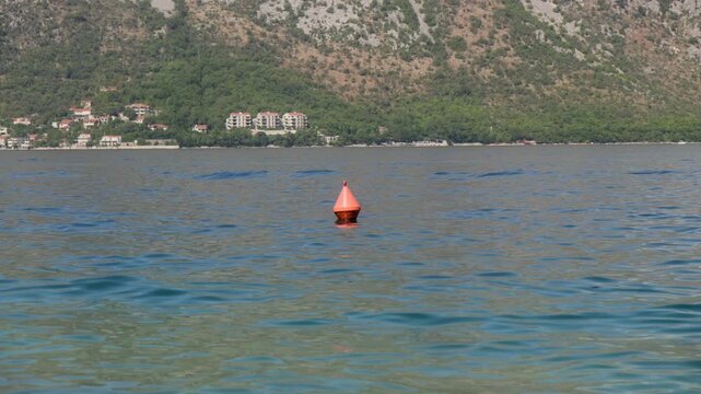 A bright orange conical buoy bobs on calm blue water as small waves shimmer. Red roof Mediterranean buildings sit below rocky tree covered slopes in midday light.