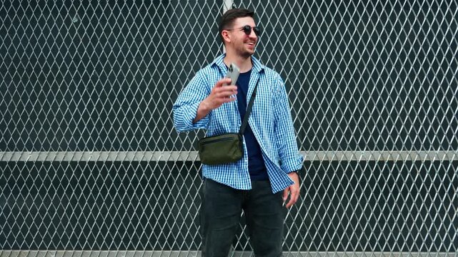 Man wearing sunglasses stands near a chain link fence on a sunny day in an urban area