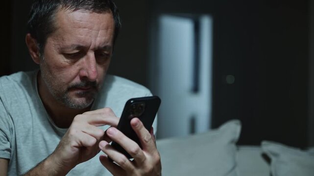 Tired mature man typing text message on smartphone in dark living room interior, using mobile phone with index finger, low key portrait in casual home setting. Selective focus. 4K.