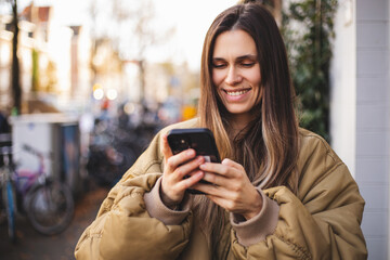 Fototapeta premium Happy brunette woman in stylish bomber jacket smiling while looking at smartphone outdoors, enjoying social media, texting, and modern urban lifestyle