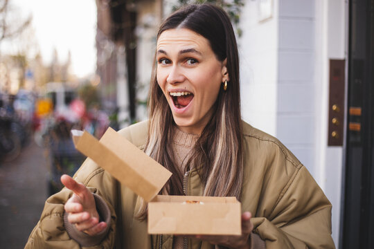 Happy brunette woman holding open dessert box with cream-filled Danish pastry, amazed and joyful, showing wow expression, enjoying sweet treat, indulgence, bakery delight, urban casual lifestyle