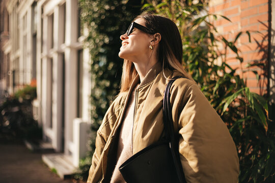 Side view of a happy brunette woman wearing sunglasses and gold bomber jacket outdoors on a sunny day, smiling joyfully, cheerful and confident, stylish, modern street style, urban lifestyle portrait.
