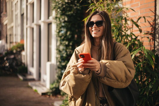 Portrait of happy brunette woman wearing glasses texting on smartphone, smiling and laughing after receiving good news, positive emotions, modern communication, urban lifestyle, stylish and confident