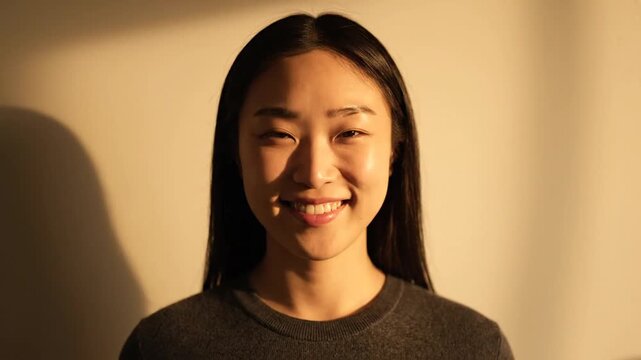 A young Asian woman smiles genuinely for 8 seconds, her expression subtly transitioning from a soft smirk to a wide, happy grin, captured in a warm, cinematic light against a shaded wall.