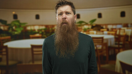 Obraz premium Man with long beard scrunches nose in dimly lit restaurant filled with round tables and chairs; disgust.