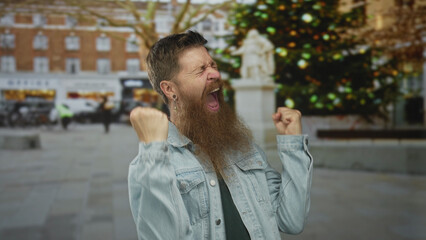Naklejka premium Young caucasian bearded man with fists raised and mouth open shouting on busy urban street at daytime; celebration.
