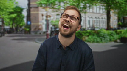 Fototapeta premium Man singing with open mouth on a city street in front of building facade, showing teeth and wearing glasses and navy shirt; joy celebration.