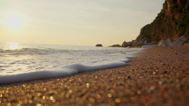 Small foamy waves lap pebbly shore at golden hour, Montenegro. Rocky cliff and boulders at right, sun reflection at left, warm backlighting and strong bokeh.