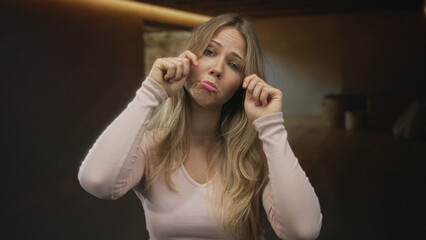 Fototapeta premium Blonde woman wearing pink top wipes tears at her eyes beside wooden table under soft warm light in room; sadness sorrow vulnerability.
